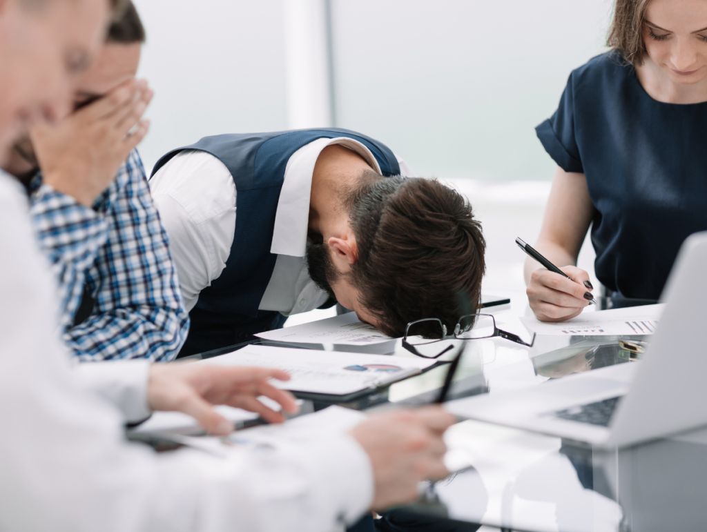 Un chef d'entreprise épuisé, la tête sur la table, entouré de collègues stressés en pleine réunion, illustrant une situation de crise et d’urgence en communication.